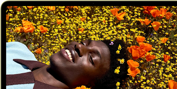 MacBook Neo amarelo-cítrico com a tela mostrando uma pessoa sorrindo deitada em um campo de flores ensolarado