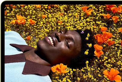 MacBook Neo amarelo-cítrico com a tela mostrando uma pessoa sorrindo deitada em um campo de flores ensolarado