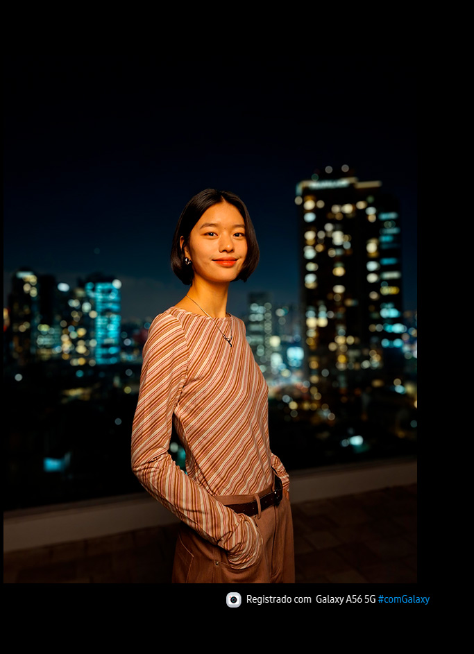 Vemos uma pessoa sorrindo para a c&acirc;mera e a vista noturna de uma cidade ao fundo. Na parte inferior, h&aacute; um &iacute;cone de c&acirc;mera com o texto "Capturado com Galaxy A56 5G #withGalaxy".